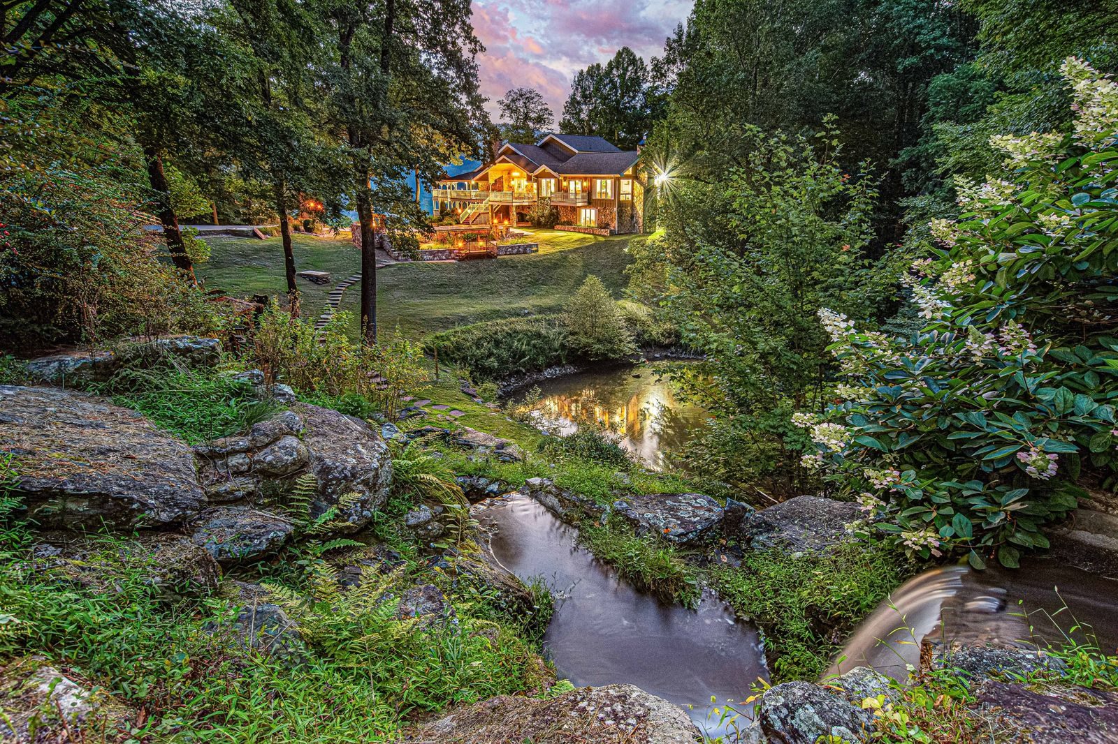 Creek and stone garden below a lit mountain home at dusk